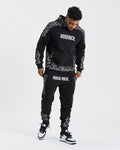 A male model wearing a black joggers with white motif print and text 'HOODRICH' on the hoodie.