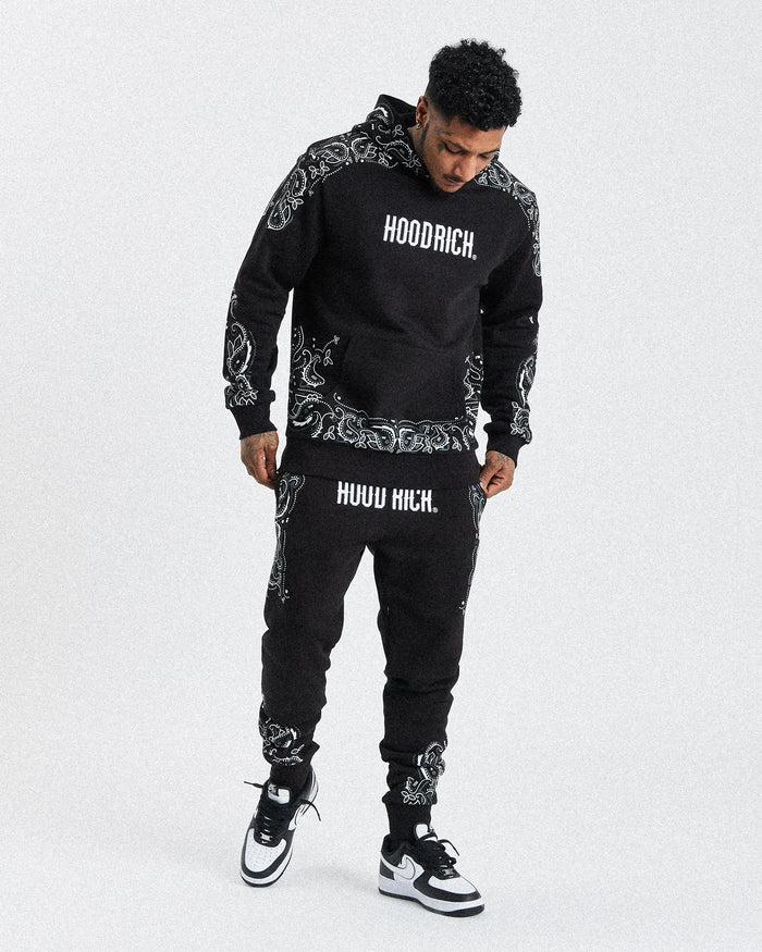 A male model wearing a black joggers with white motif print and text 'HOODRICH' on the hoodie.