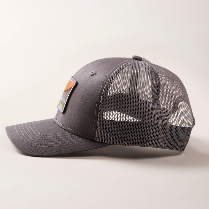 The Mountain Trucker Hat- Charcoal Grey features a curved brim, mesh back panels, and a colorful rectangular front patch. Displayed on a light background, it has an adjustable snap back for a comfortable fit.