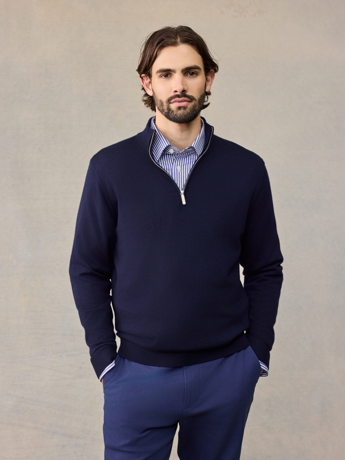 Man in navy quarter-zip sweater over striped collared shirt, navy pants, hands in pockets