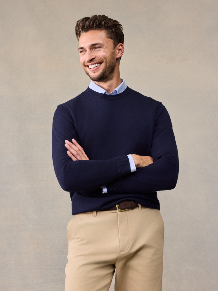 Smiling man in navy crewneck sweater over light blue collared shirt, arms crossed, khaki pants