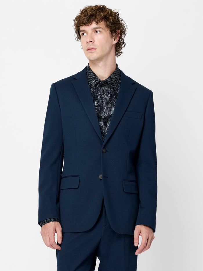 A man wearing a navy Piqué Suit Jacket  with long sleeves, a chest welt pocket and 2 front pockets.