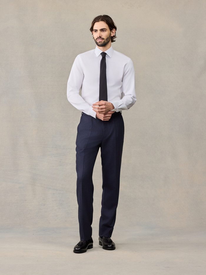 Man in navy plaid wool suit pants and white shirt with black tie, hands clasped, full length