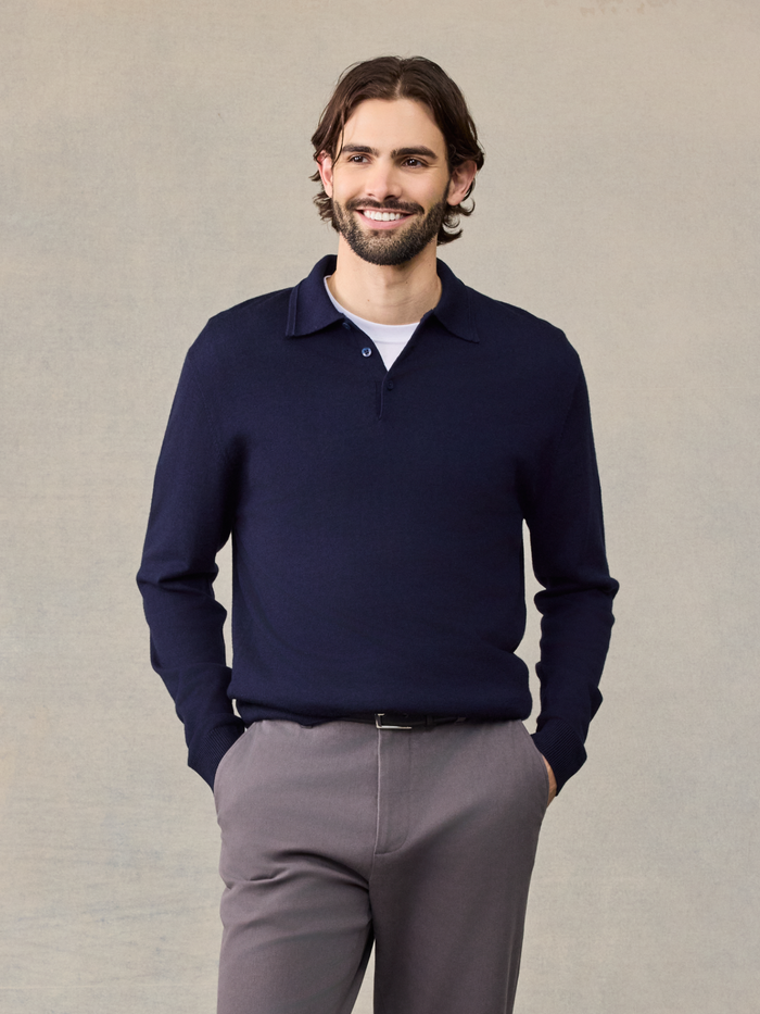 Smiling man in navy long-sleeve polo sweater over white tee, grey pants, hands in pockets