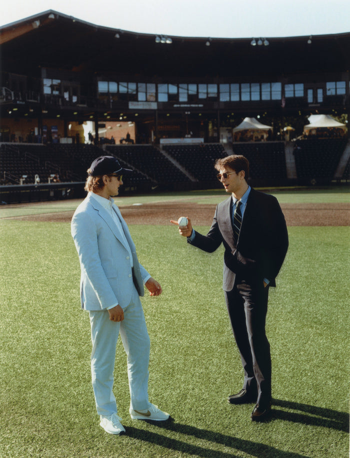 Two men on a baseball field, one in a navy seersucker suit, the other in a dark suit holding a baseball
