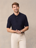 Smiling man in navy short-sleeve sweater polo and cream pants, hands clasped