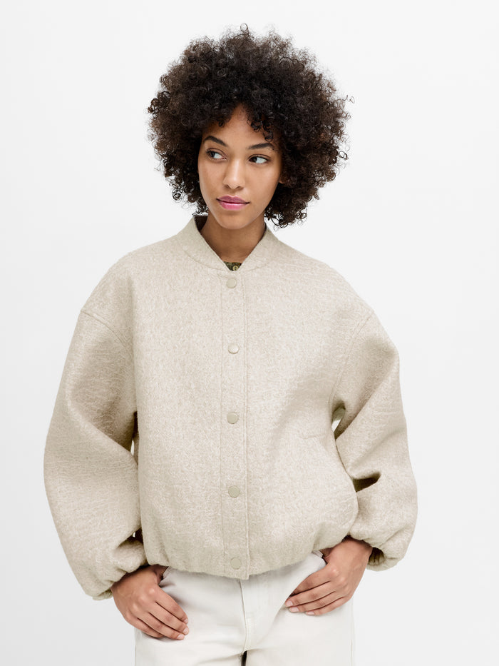 A woman wearing an oatmeal textured bomber jacket with 2 side pockets, puff sleeves and drop shoulders.