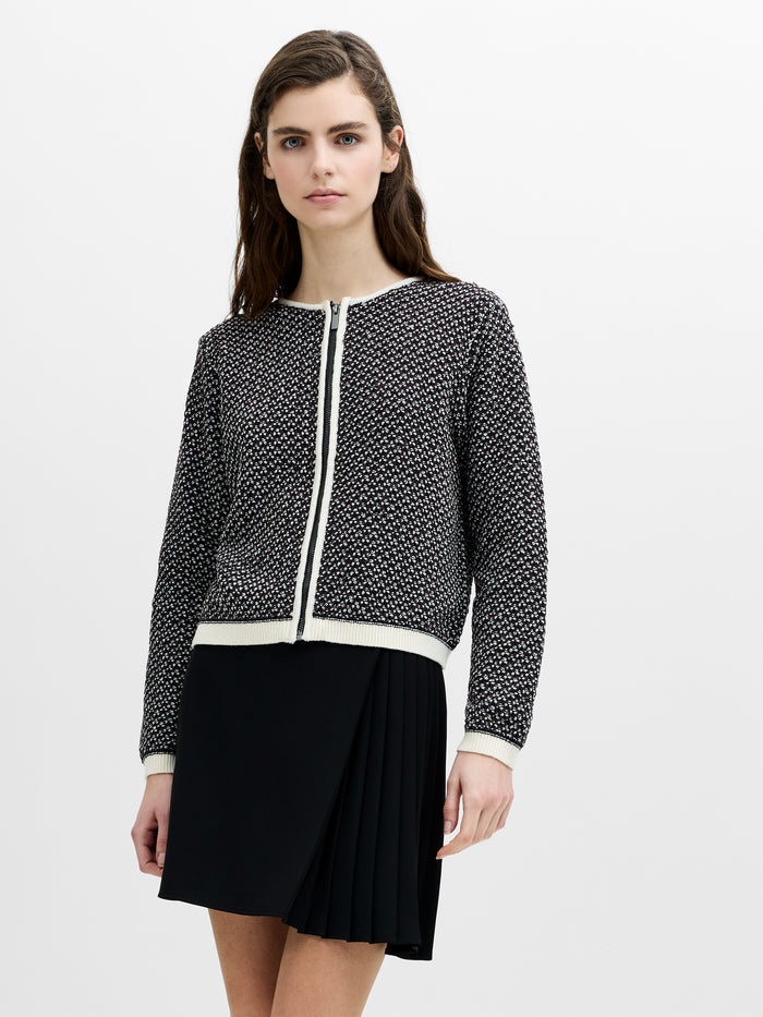 A woman wearing a black and cream zip through knit, box fit cardigan with long sleeves and a round neck.