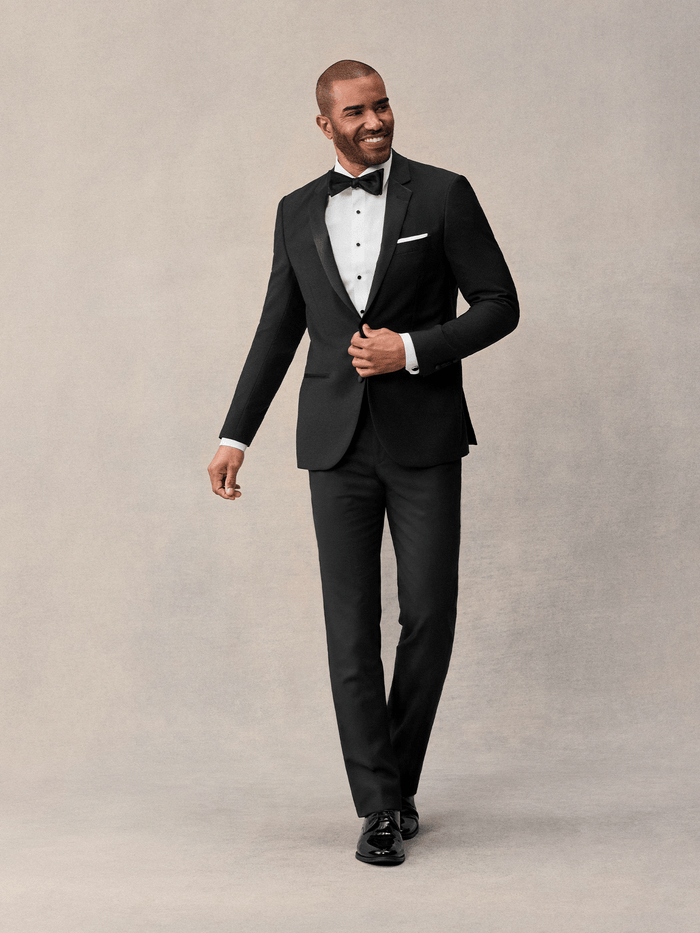 Man in black notch lapel tuxedo with bow tie and white pocket square, walking against cream backdrop