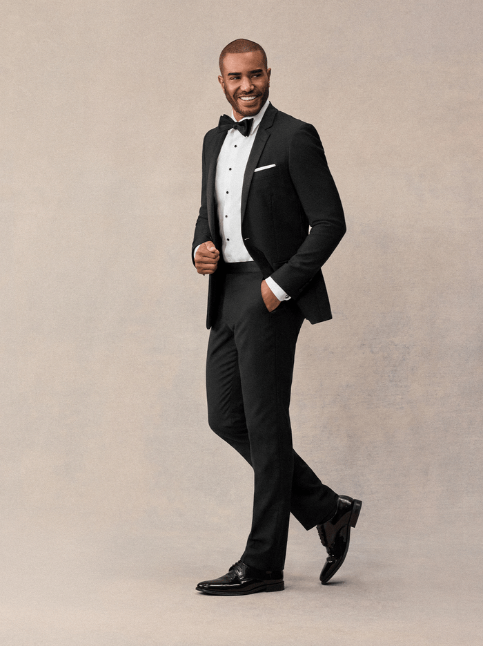 Man in black notch lapel tuxedo with bow tie, smiling, walking against cream backdrop