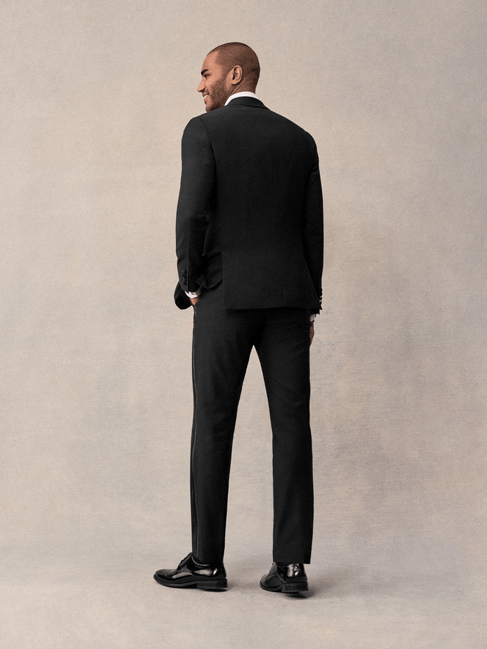 Back view of man in black notch lapel tuxedo, hand in pocket, against cream backdrop
