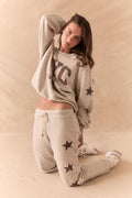 Full body view of model wearing NYC Stars Pullover in Heather Grey with smoky plum star sleeves and matching joggers, styled casually, against a beige background