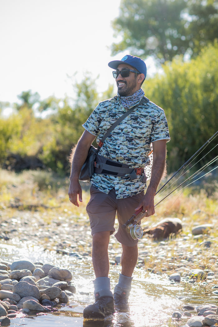 A man in a Nylon Trout Hat- Navy Blue stands on a rocky riverbank, holding fishing rods and smiling in the sunlight, surrounded by green foliage.