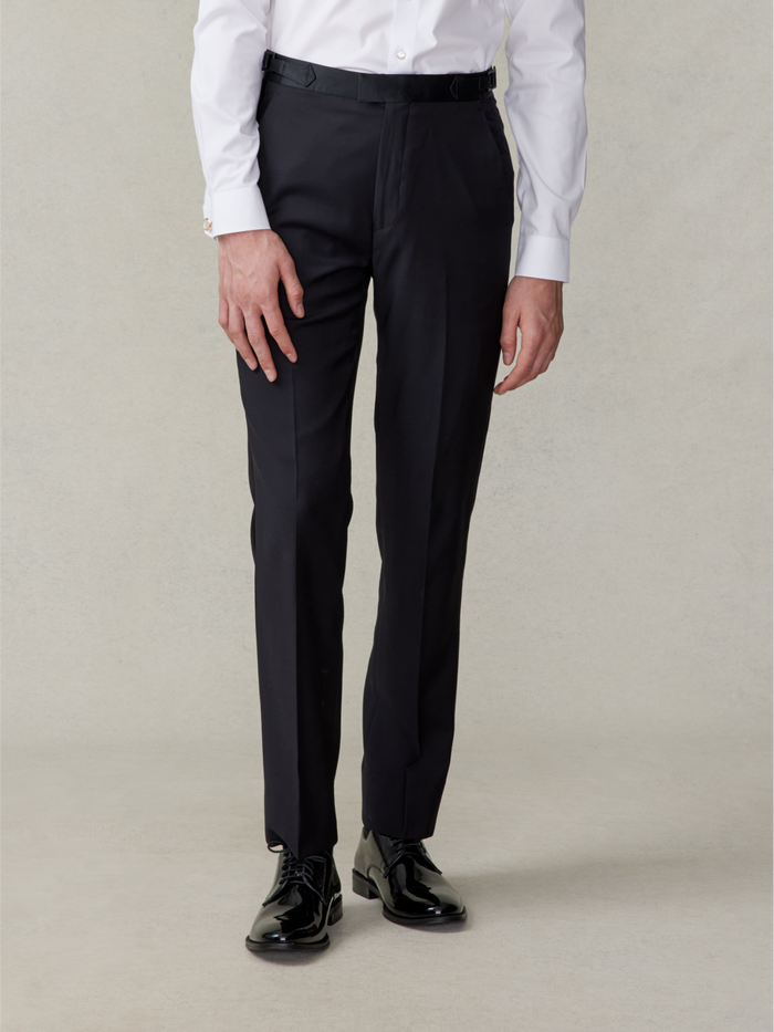 Man in black tuxedo trousers and patent shoes, front view, beige backdrop.