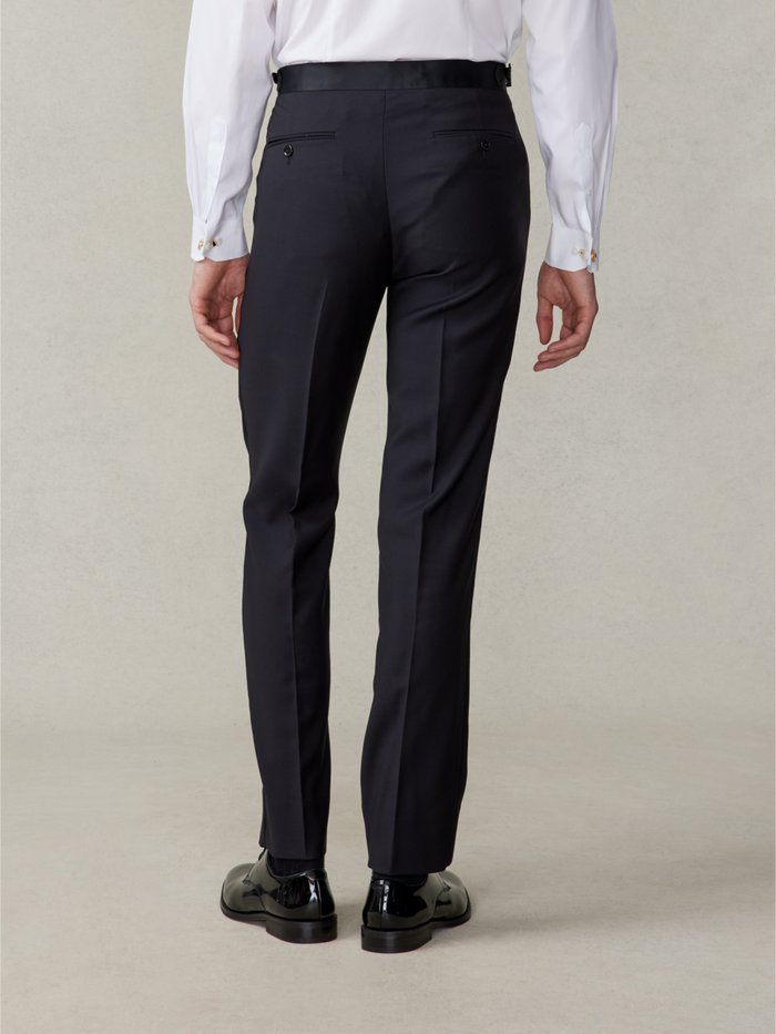 Man in black tuxedo trousers and patent shoes, back view, beige backdrop.