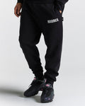 A person wearing black joggers with 'HOODRICK' branding on the left leg and white sneakers.