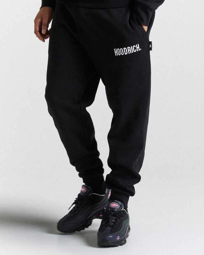 A person wearing black joggers with 'HOODRICK' branding on the left leg and white sneakers.