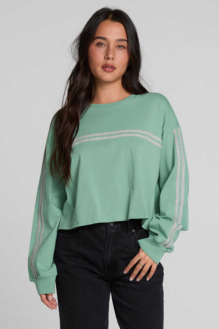 A woman modeling a sage green cropped sweatshirt with light gray stripe details on the chest and sleeves, paired with black jeans, against a solid light gray background