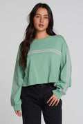 A woman modeling a sage green cropped sweatshirt with light gray stripe details on the chest and sleeves, paired with black jeans, against a solid light gray background