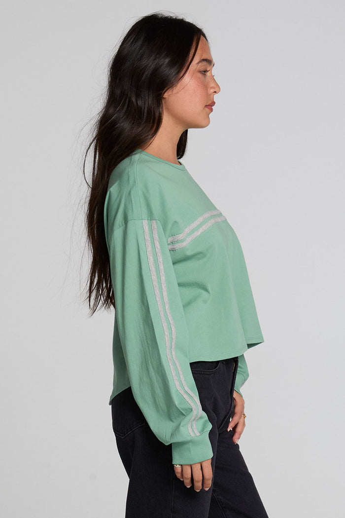 A woman in profile wearing a sage green cropped long-sleeve sweatshirt with white double stripes on the sleeves, paired with black jeans, against a light gray studio background