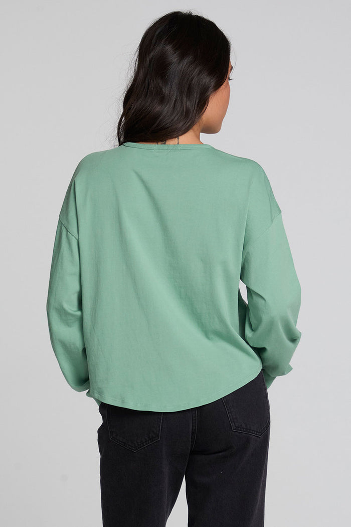 A woman viewed from the back wearing a sage green long-sleeve top and dark jeans against a light gray studio background.