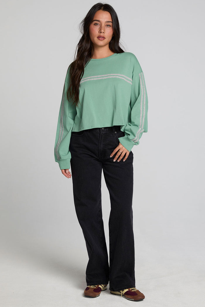 A woman modeling a sage green long-sleeve crewneck sweatshirt with thin white horizontal stripes across the chest and sleeves, paired with black wide-leg jeans and brown shoes, against a light gray studio background