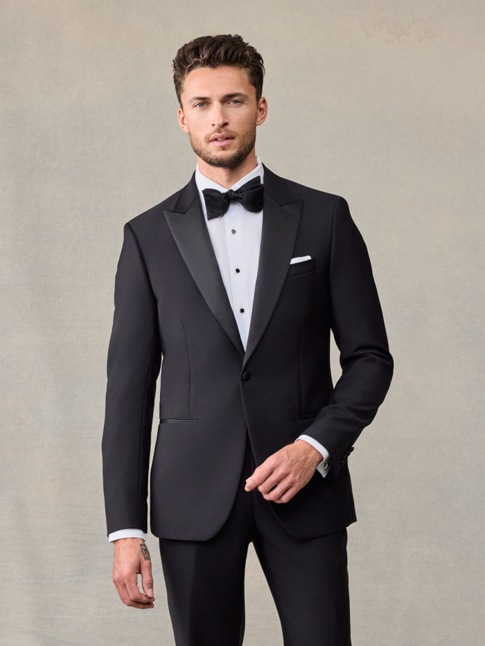 Man in black peak lapel tuxedo with bow tie and pocket square, front view, on beige backdrop