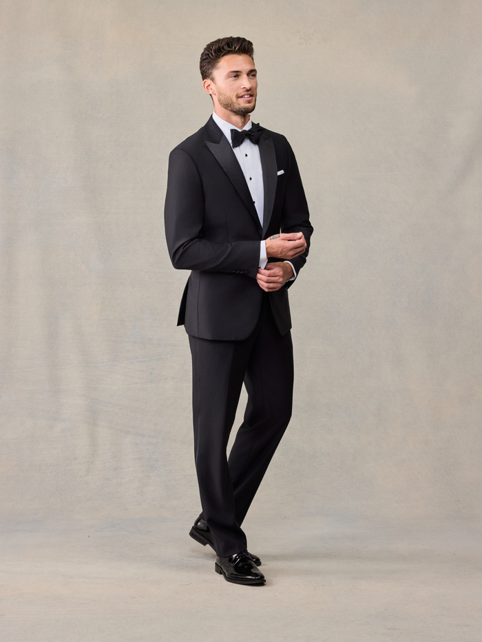 Man in black peak lapel tuxedo with bow tie, three-quarter view, smiling, on beige backdrop