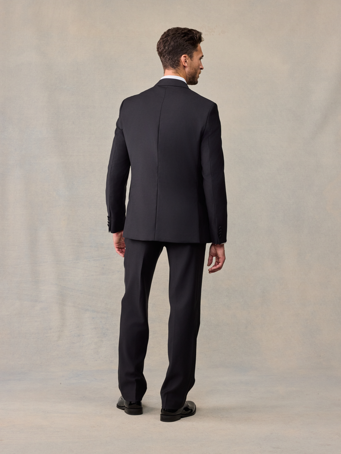 Back view of man in black peak lapel tuxedo jacket and trousers on beige backdrop