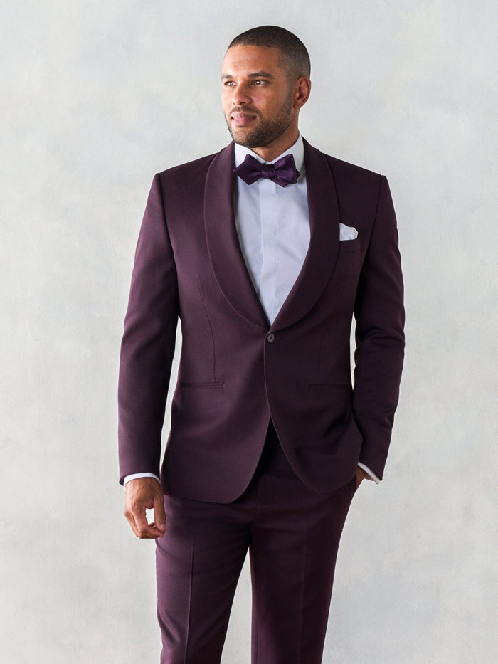 Man in plum shawl lapel tuxedo suit, hand in pocket, looking aside, grey backdrop