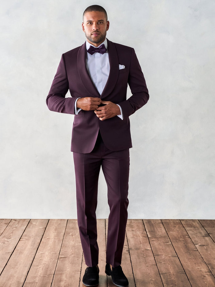 Man in plum shawl lapel tuxedo suit adjusting jacket button, matching plum bow tie, on wood floor