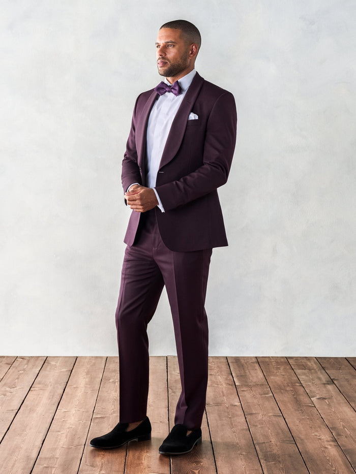 Man in plum shawl lapel tuxedo suit, three-quarter turned view, hands clasped, matching bow tie