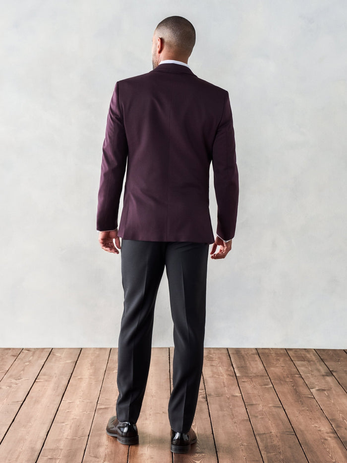 Back view of man in plum shawl lapel tuxedo jacket and black trousers on wood floor