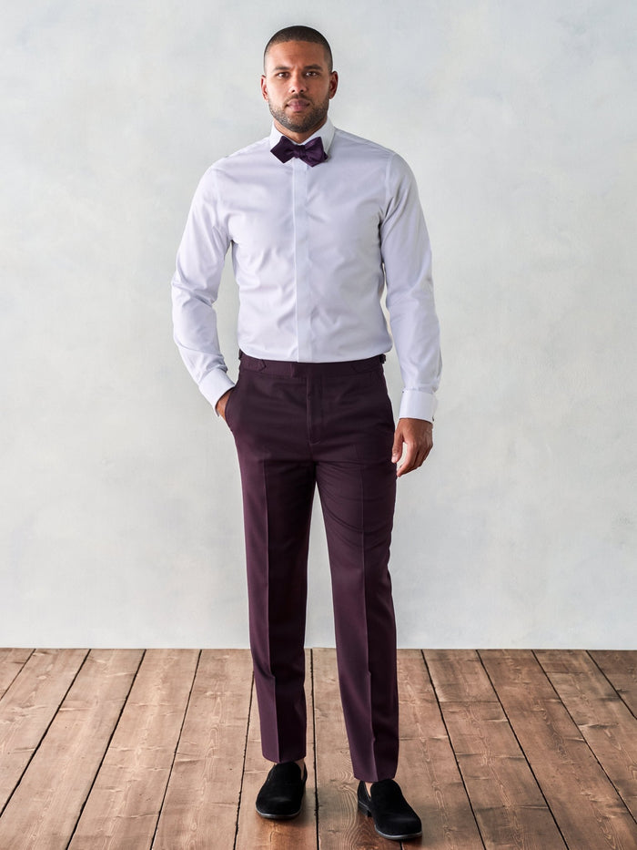 Man in white tuxedo shirt and plum trousers with matching bow tie, hands in pockets, on wood floor