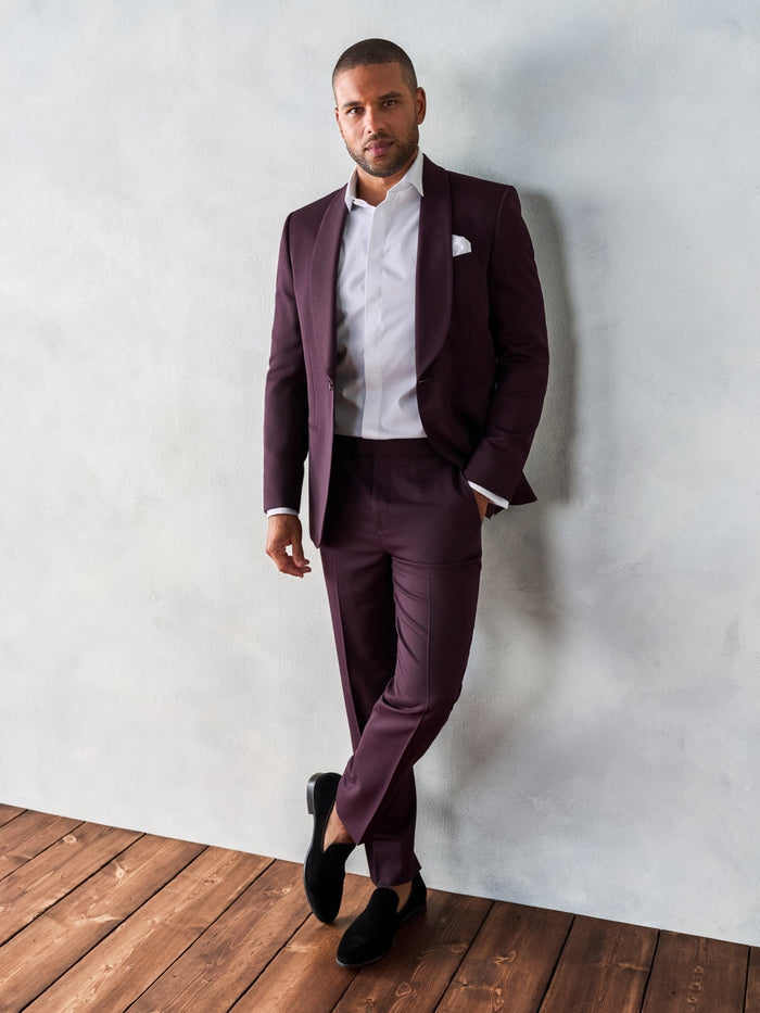 Man in plum shawl lapel tuxedo jacket leaning against wall, plum trousers, on wood floor
