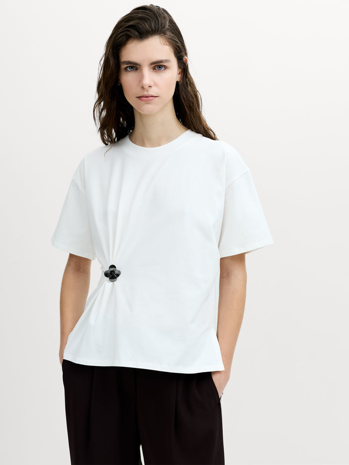 A woman wearing a white dropped shoulder short sleeved T-shirt, with a crew neck and a removable metal floral detail for a cinched fit.
