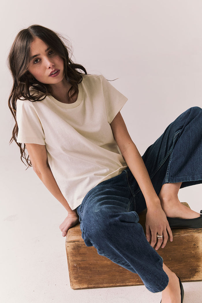 Woman sitting on a wooden box wearing the Rambler Starry White Tee with ringer neck and cap sleeves and blue jeans against a plain background