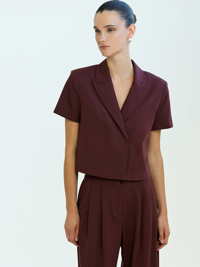 A woman wearing a burgundy padded shoulder Blazer with short sleeves, a v neckline, a split lapel collar, and a cropped box fit.