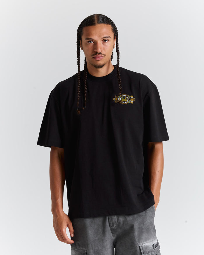 A man with long braids wearing a black crewneck t-shirt with a small gold and green graphic on the left chest, paired with gray cargo pants, against a light gray background.