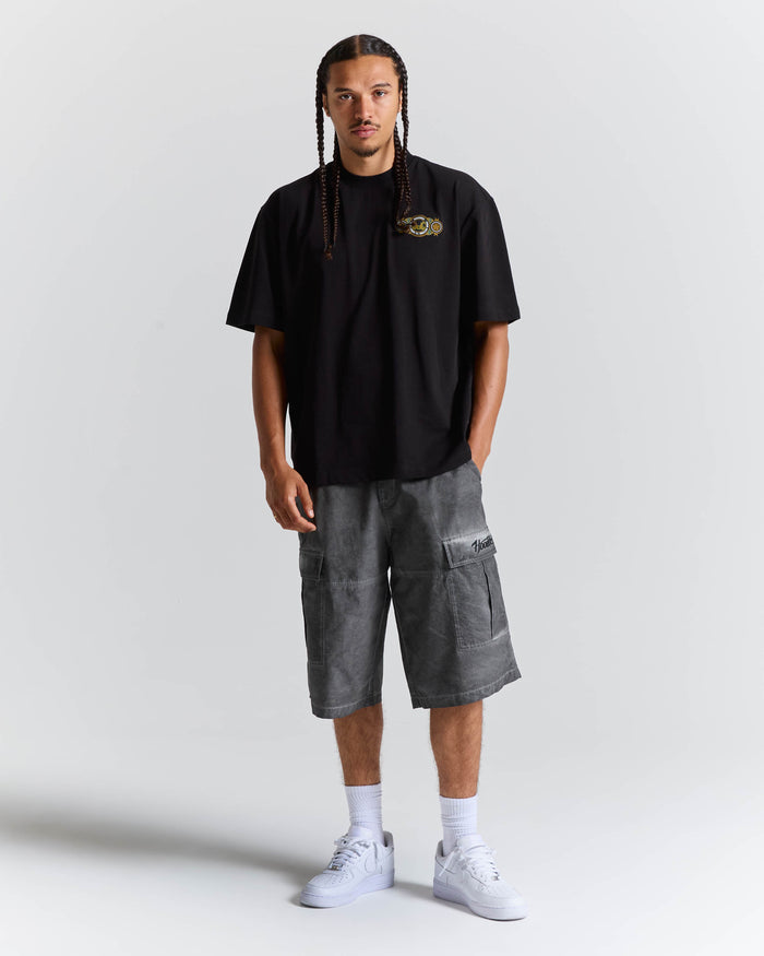 A man modeling a black short-sleeve t-shirt with a small gold and green chest graphic, paired with gray washed cargo shorts, white socks, and white sneakers, standing against a light gray studio background.