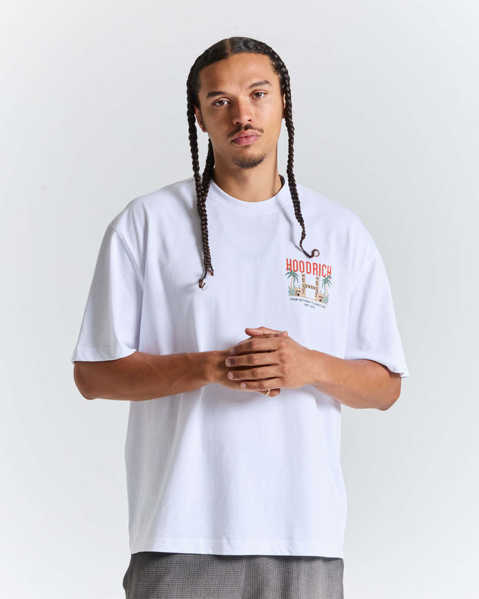 A man with braided hair wearing a white short-sleeve t-shirt with a colorful 'HOODRICH' graphic on the left chest, posed against a light gray studio background.