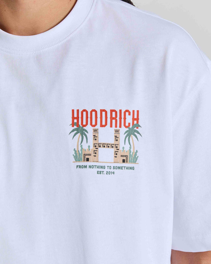 A close-up of a person wearing a white crewneck t-shirt with a colorful graphic on the chest. The graphic features the word 'HOODRICH' in red-orange letters above a beige building flanked by green palm trees, with the text 'FROM NOTHING TO SOMETHING EST.