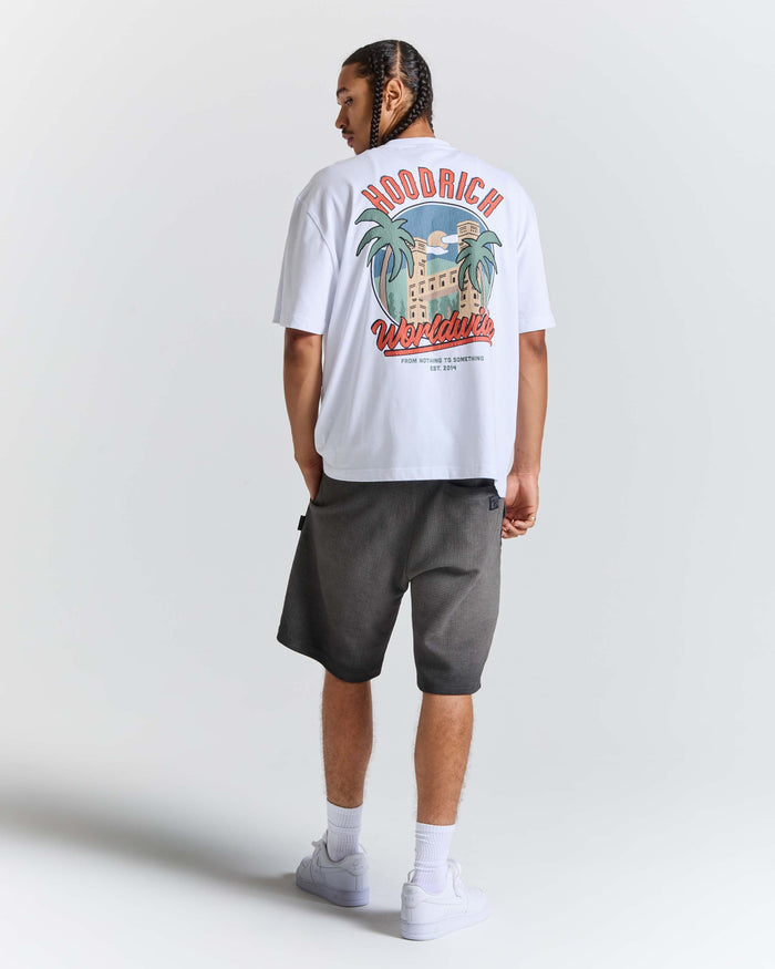 A man wearing a white HODRICH t-shirt with a large colorful graphic on the back, paired with dark gray shorts, white socks, and white sneakers, shown from a rear three-quarter view against a light gray studio background.