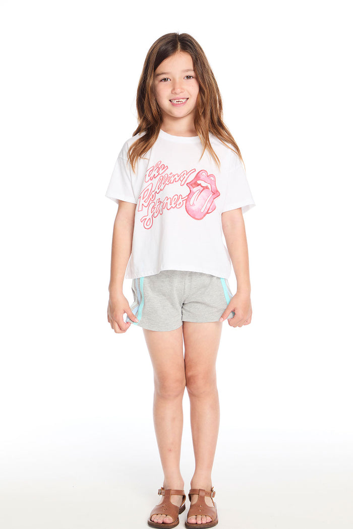 Girl wearing the Chaser Kids Rolling Stones Painterly Tongue Logo Tee paired with grey shorts featuring rainbow side stripes and tan sandals on a white background
