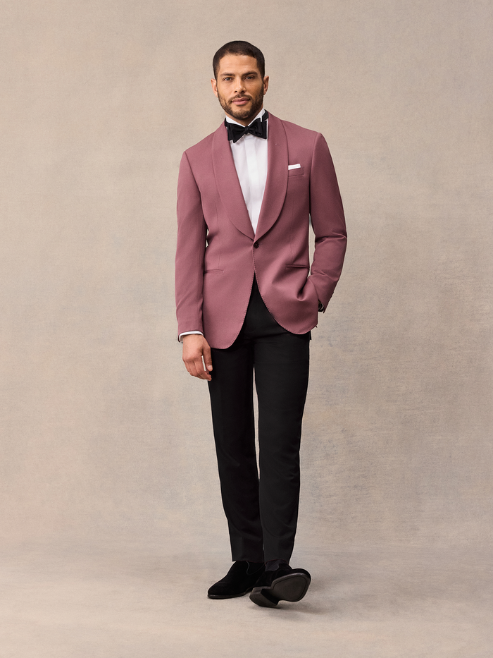 Man in dusty rose shawl collar tuxedo jacket and black trousers with black bow tie, front view, on beige backdrop
