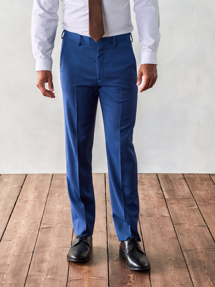 Man in royal blue suit pants, white shirt, and brown tie, front view on wood floor