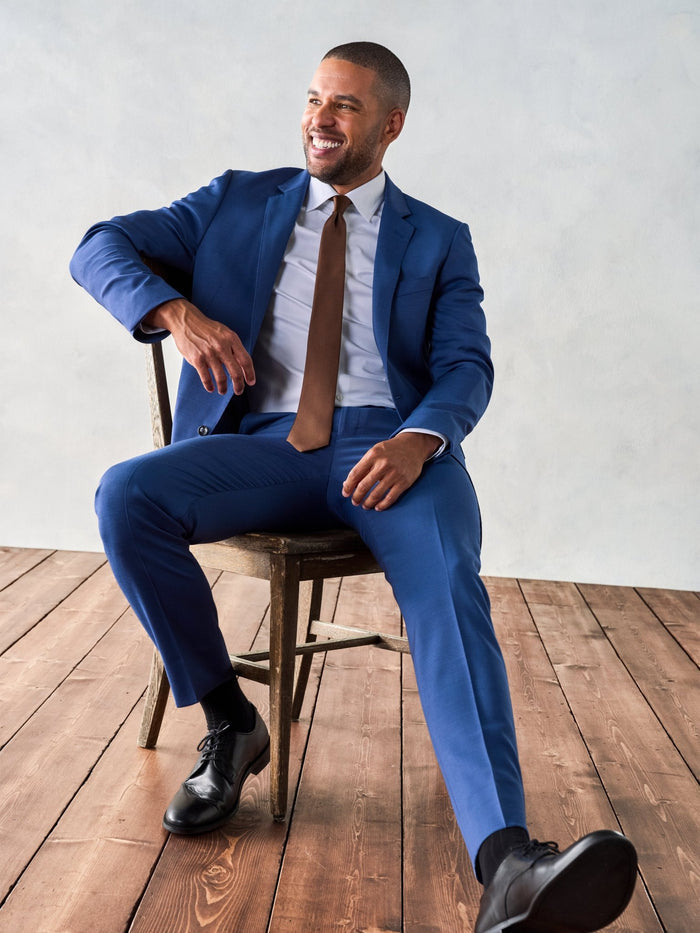 Man in royal blue suit and brown tie, seated on chair, smiling