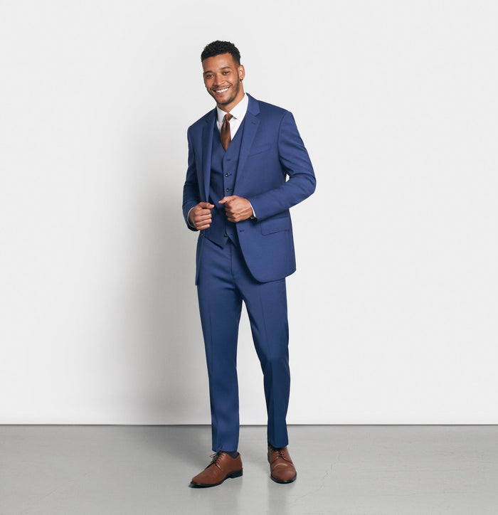 Man in royal blue three-piece suit with brown tie and shoes, buttoning jacket, smiling
