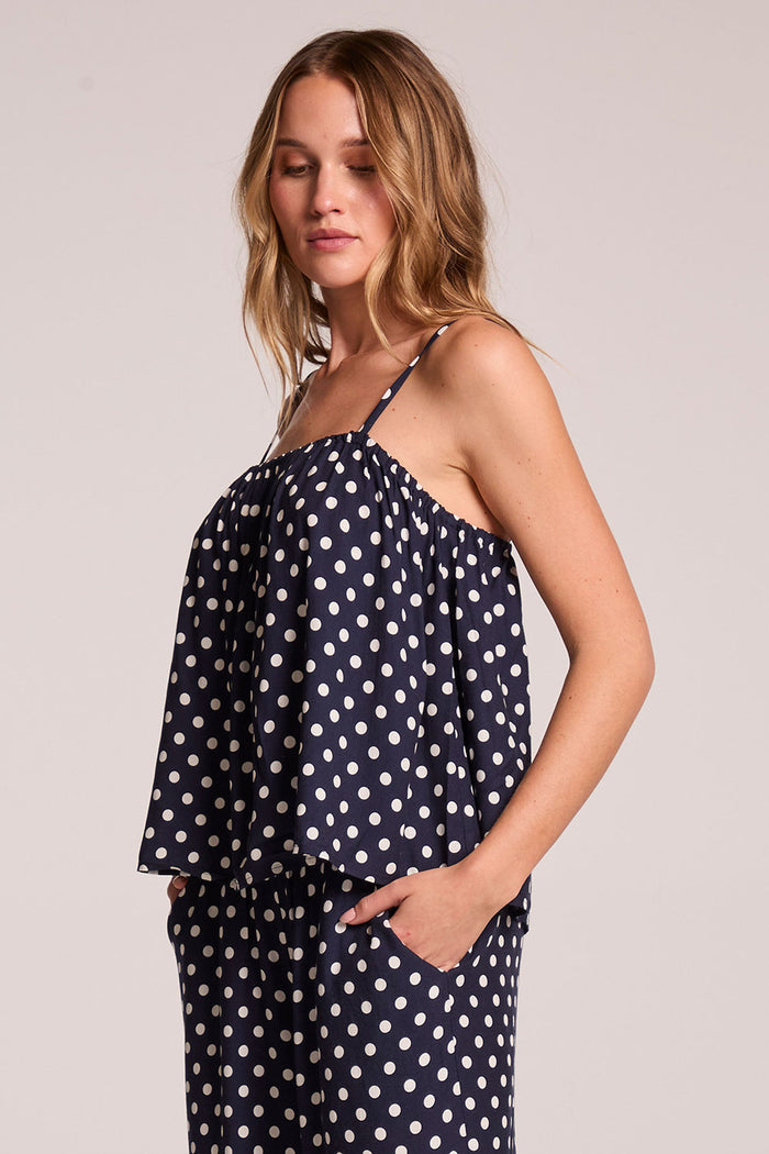 Side angle view of a woman wearing the Chaser   Rubie Navy Spot Tank Top on a neutral background with hands in her pockets