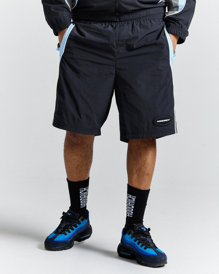 Person wearing black shorts with a brand logo, black socks, and blue sneakers on a light grey background